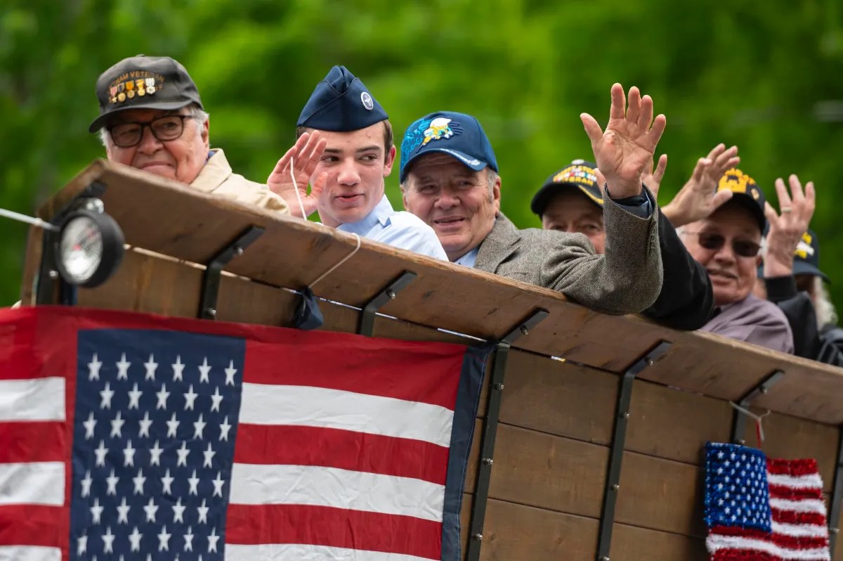 Group of men, including veterans and a uniformed service member, wave from a parade float decorated with American flags.