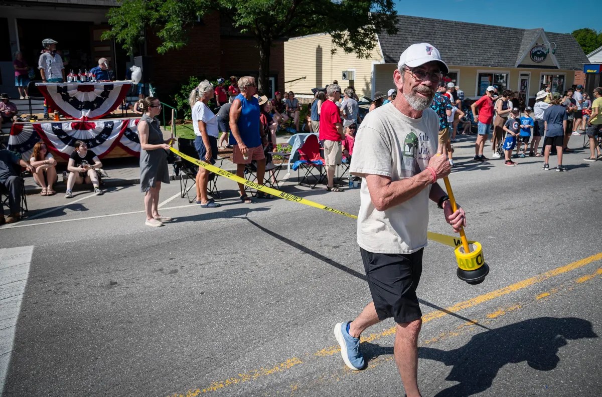 A man in a white T-shirt and black shorts walks on a street holding a plunger during a parade. Spectators are seated and standing along the sidewalk, with some decorations visible in the background.