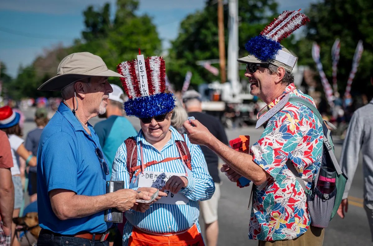 Three men, one in a blue shirt and two wearing festive hats with red, white, and blue decorations, are talking at an outdoor event. Other attendees and trees are visible in the background.