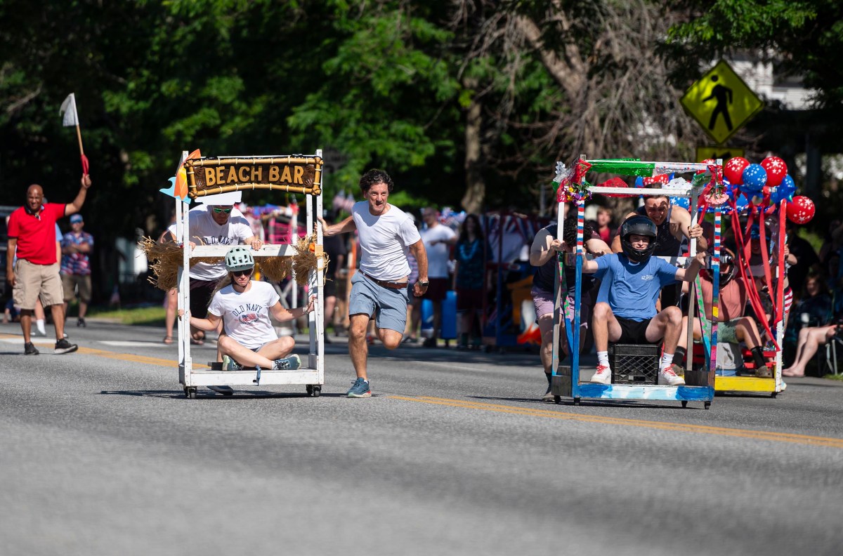 Participants in a soapbox race compete in creatively designed carts on a sunny day, with spectators watching and greenery in the background.