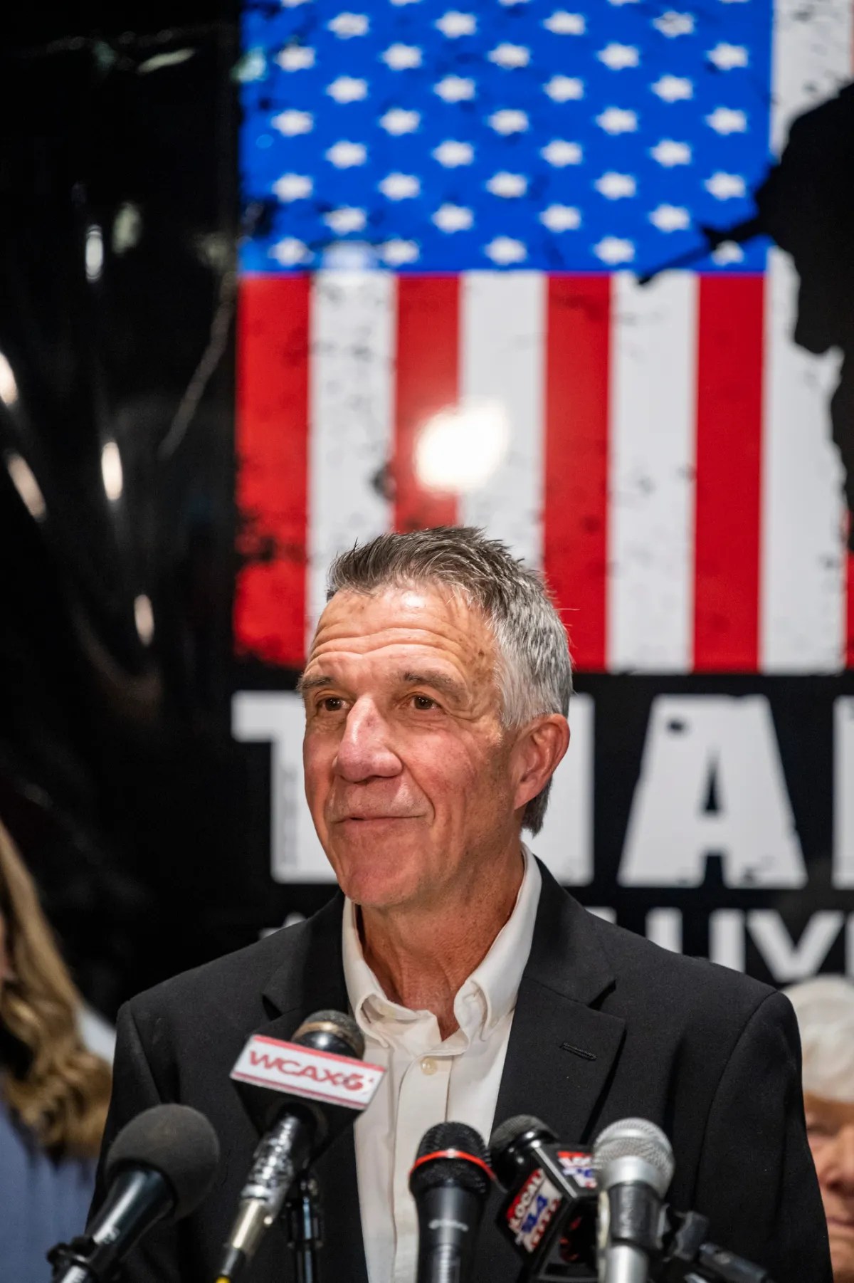 A man in a suit speaks at a podium with multiple microphones. An American flag is visible in the background.