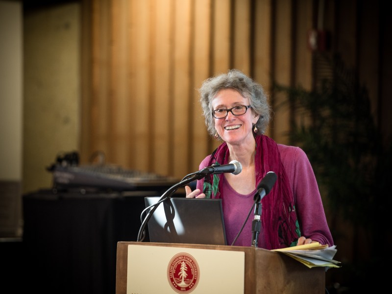 A woman with gray hair and glasses speaks at a podium with a laptop and microphone, smiling, in an indoor setting with wood paneling and audio equipment in the background.
