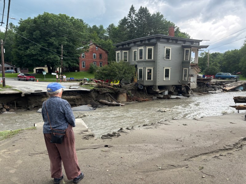 A person stands watching a house partially collapsed into a flooded and damaged road, with debris and other affected buildings visible in the background.