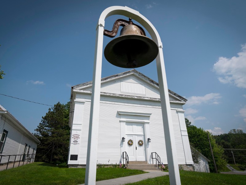 A large bronze bell mounted on a white frame stands in front of a white wooden church building with double doors and wreaths.