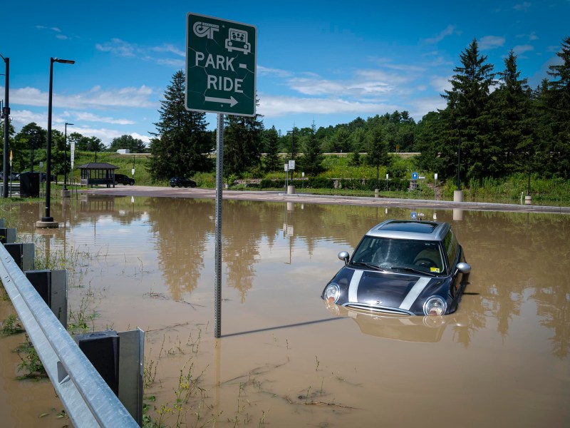 A car partially submerged in floodwater in a parking lot with a "Park and Ride" sign visible.