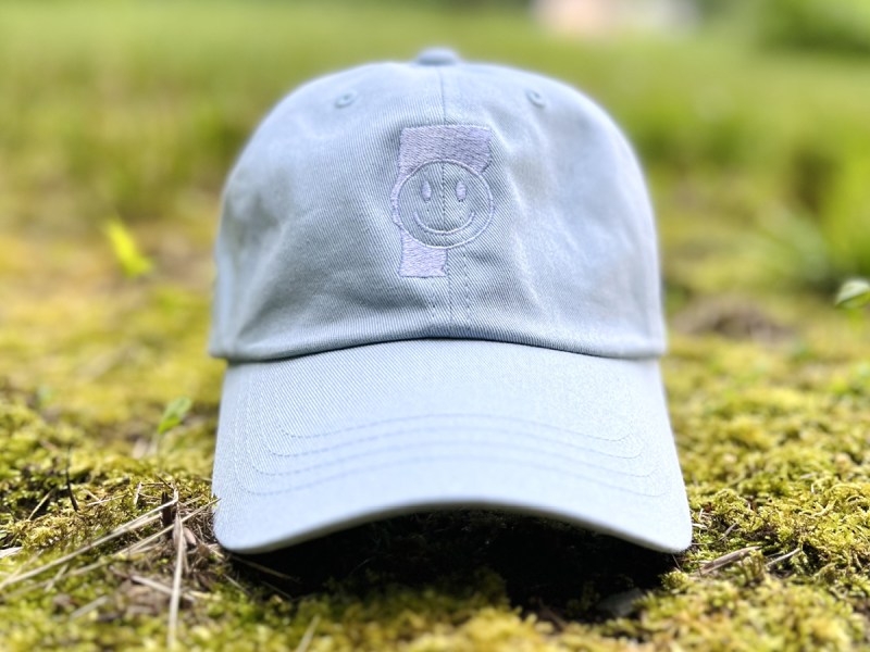 A light blue baseball cap with a smiley face patch on the front resting on mossy ground outdoors.