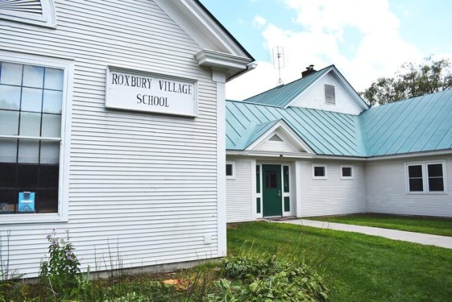 A white building with a teal metal roof and a sign reading "Roxbury Village School" on the wall. The school has multiple windows and a small garden area in front.