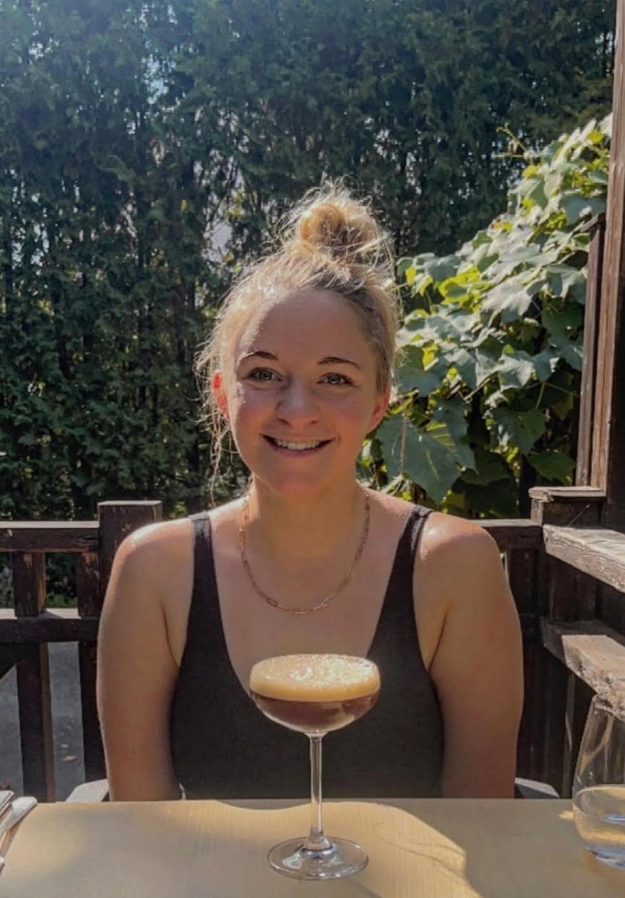 A woman sits at an outdoor table in sunlight, smiling at the camera with a cocktail in a coupe glass in front of her. Green foliage is visible in the background.