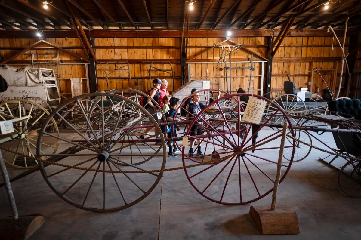 A group of people walk through a barn-like museum displaying old wooden and metal wagon wheels and vintage farm equipment.