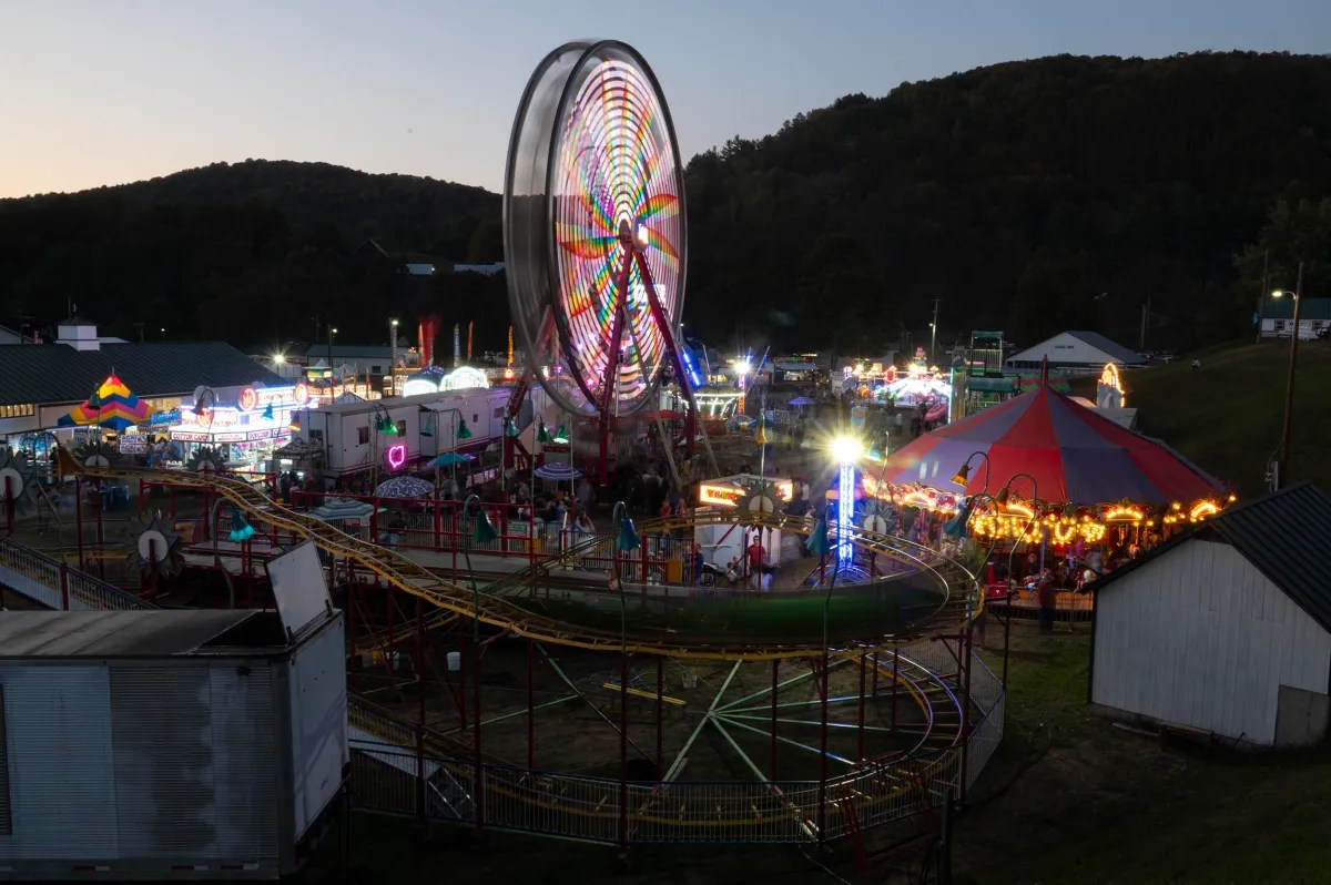 A brightly lit ferris wheel spins at a fairground at dusk, surrounded by rides, tents, and booths, with hills in the background.