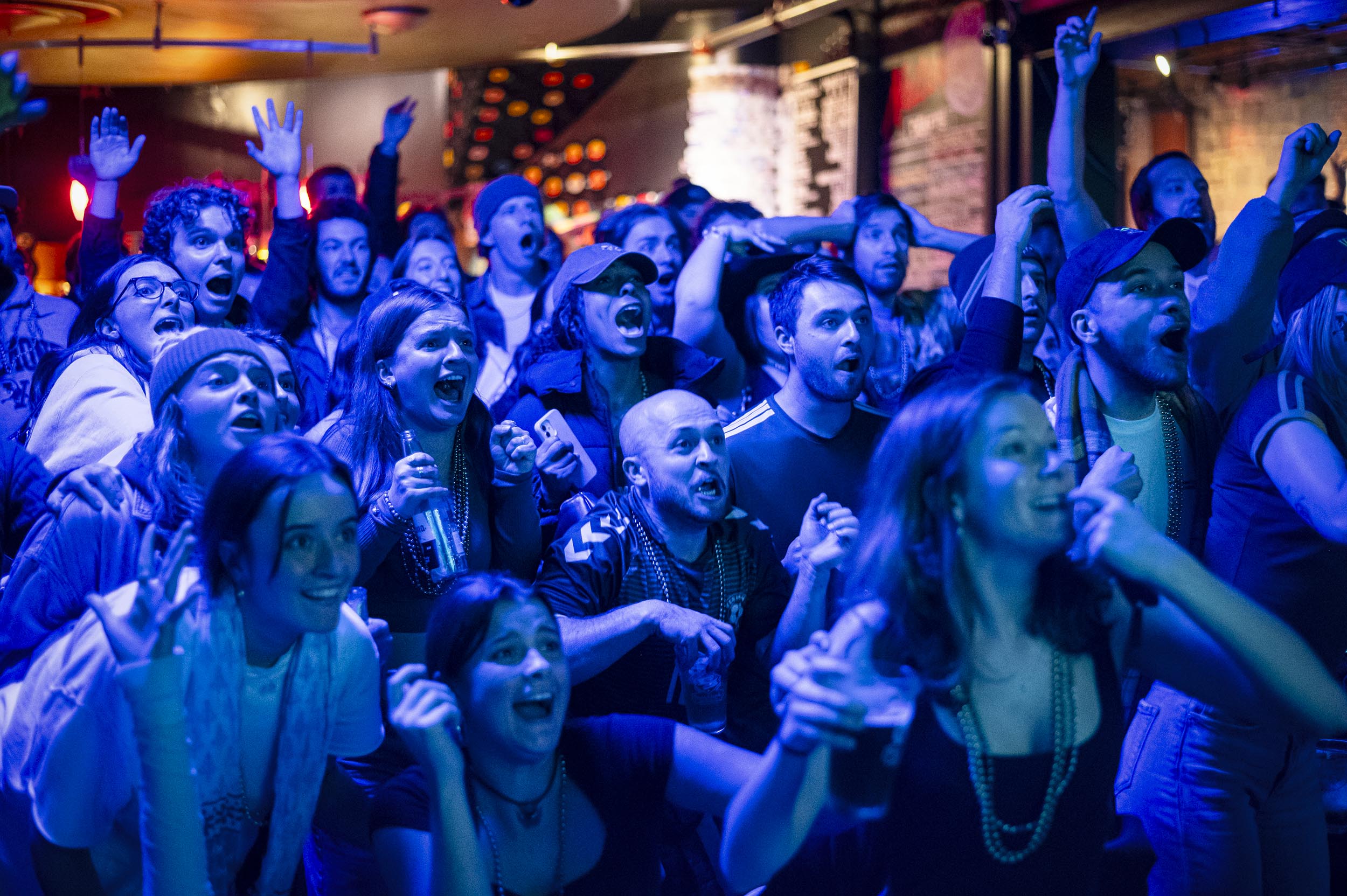 A lively crowd in a dimly lit venue cheers enthusiastically, illuminated by blue lighting.