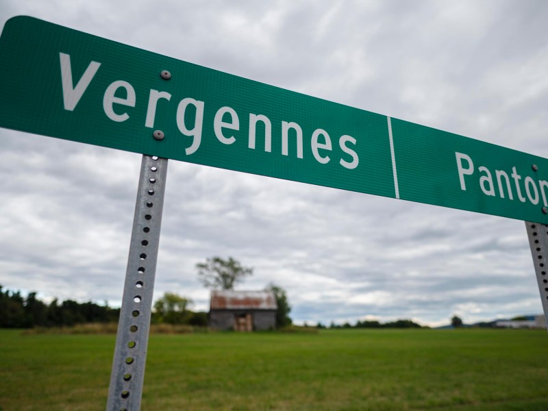 A green road sign with white text showing directions to Vergennes and Panton stands in front of a grassy field and an overcast sky.
