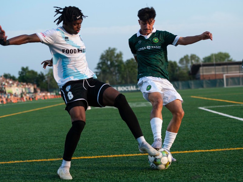 Two soccer players compete for the ball near the sideline during a match on a green field, with spectators visible in the background.