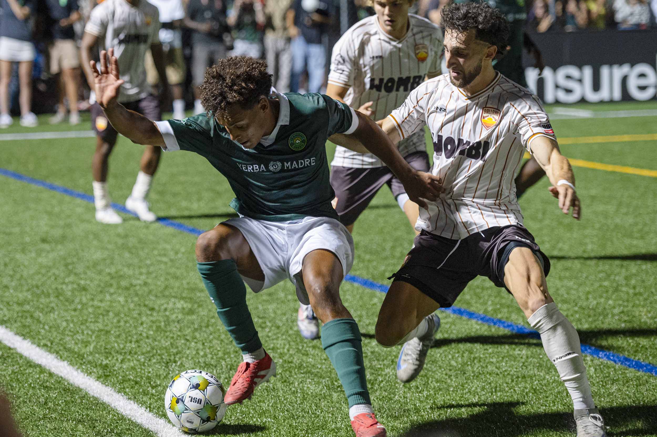 Two soccer players compete for the ball during a match on a turf field, with spectators watching in the background.