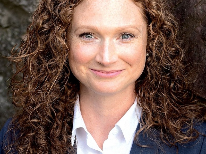 A woman with curly brown hair wearing a navy blazer and white shirt stands in front of a stone wall, smiling at the camera.