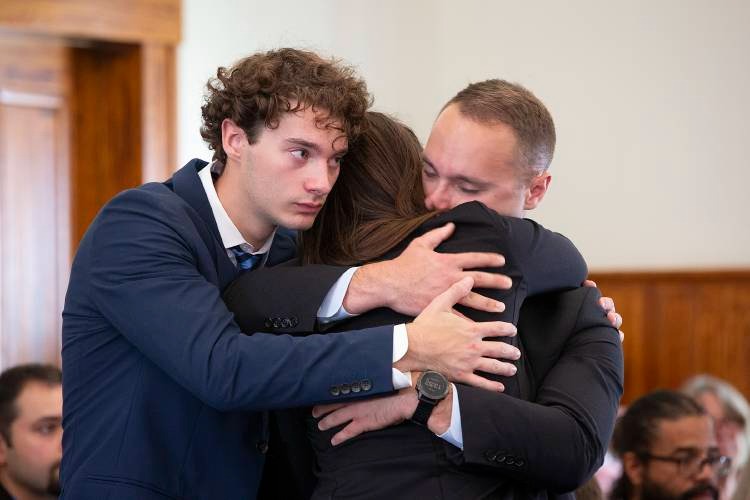 Three people dressed formally embrace each other in a courtroom setting, appearing emotional. Other individuals are seated in the background.