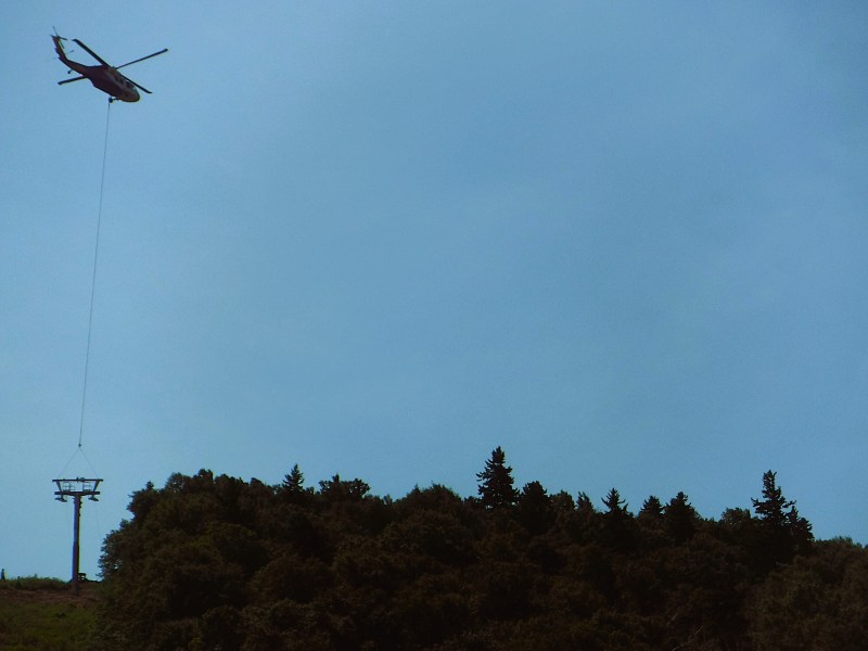 A helicopter flies above trees, carrying equipment on a cable near a ski lift tower under a clear sky.