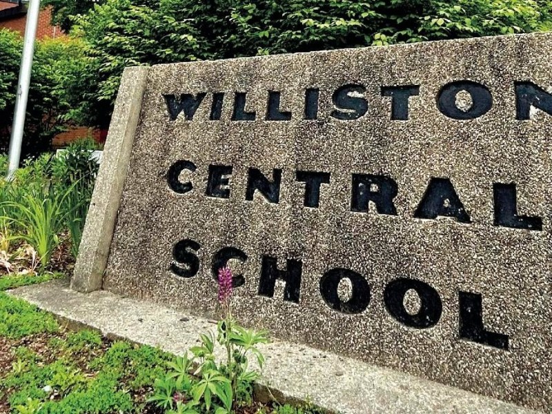 A concrete sign with "Williston Central School" in black letters stands next to greenery and plants.