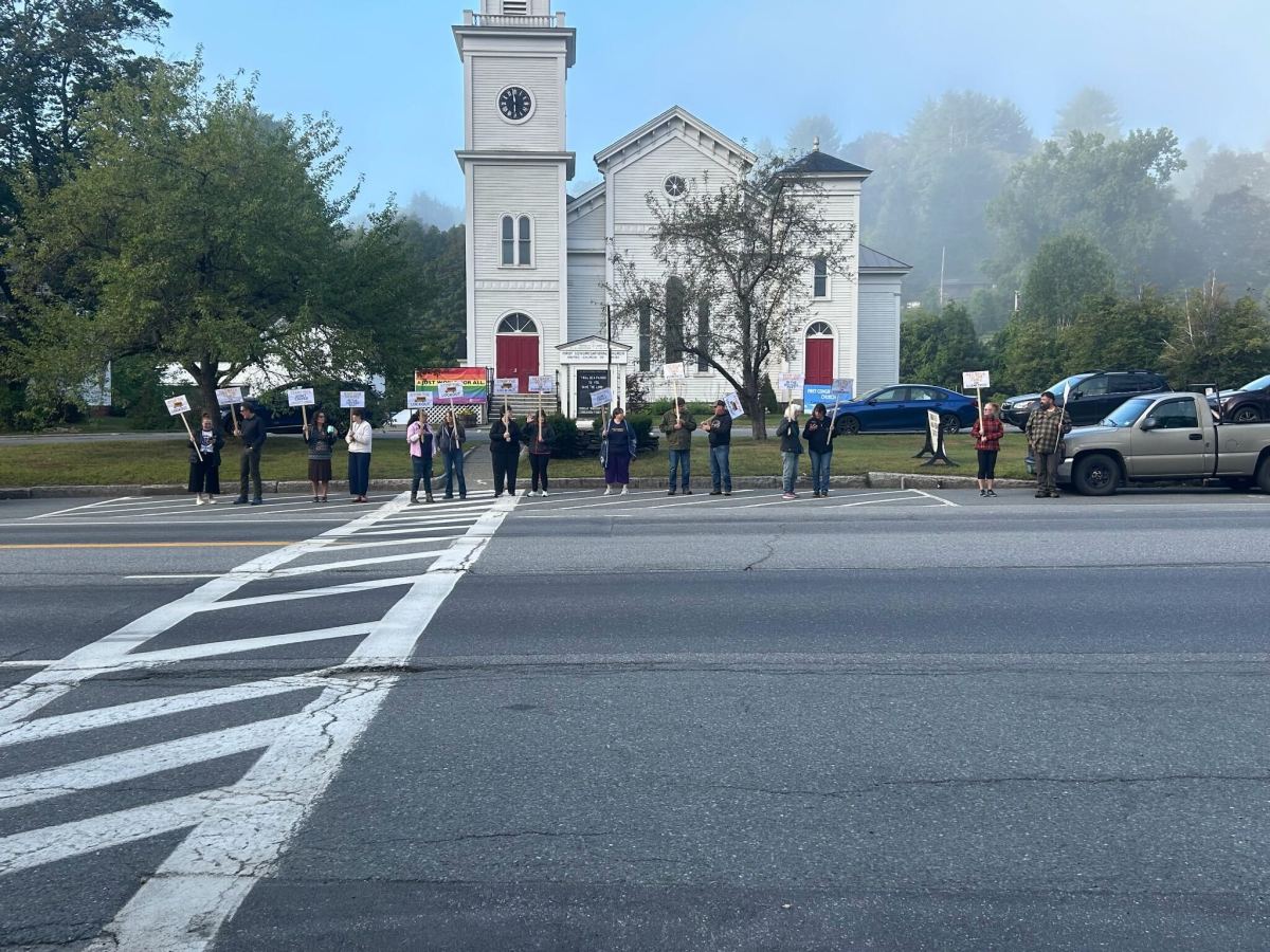 A group of people stand in a row on the roadside holding signs in front of a white church building on a foggy day.