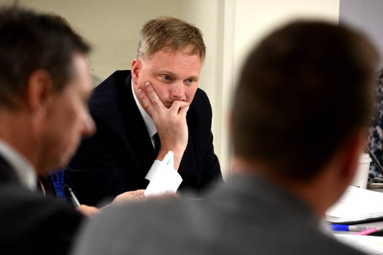 A man in a suit rests his face on his hand, appearing thoughtful, while sitting at a table with two other people during a meeting.