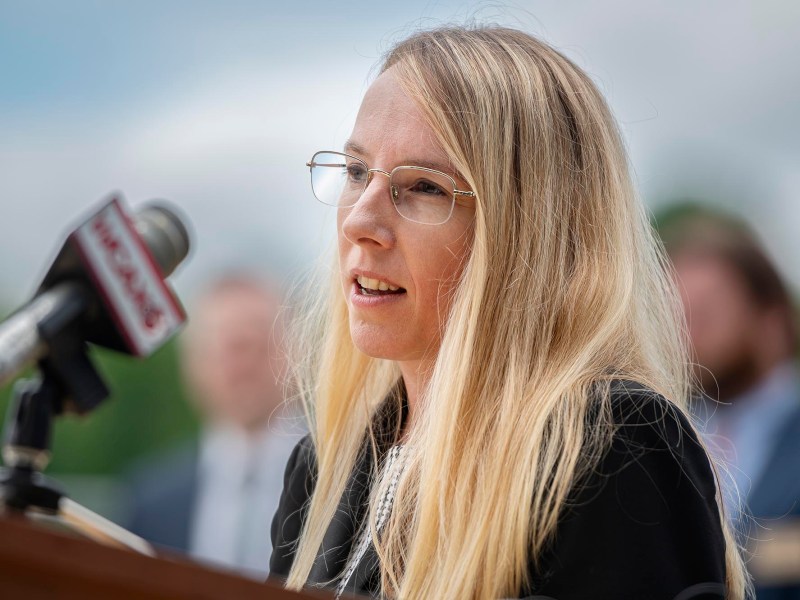 A person with long blonde hair and glasses speaks into a microphone at an outdoor event. A blurred person and green foliage are visible in the background.