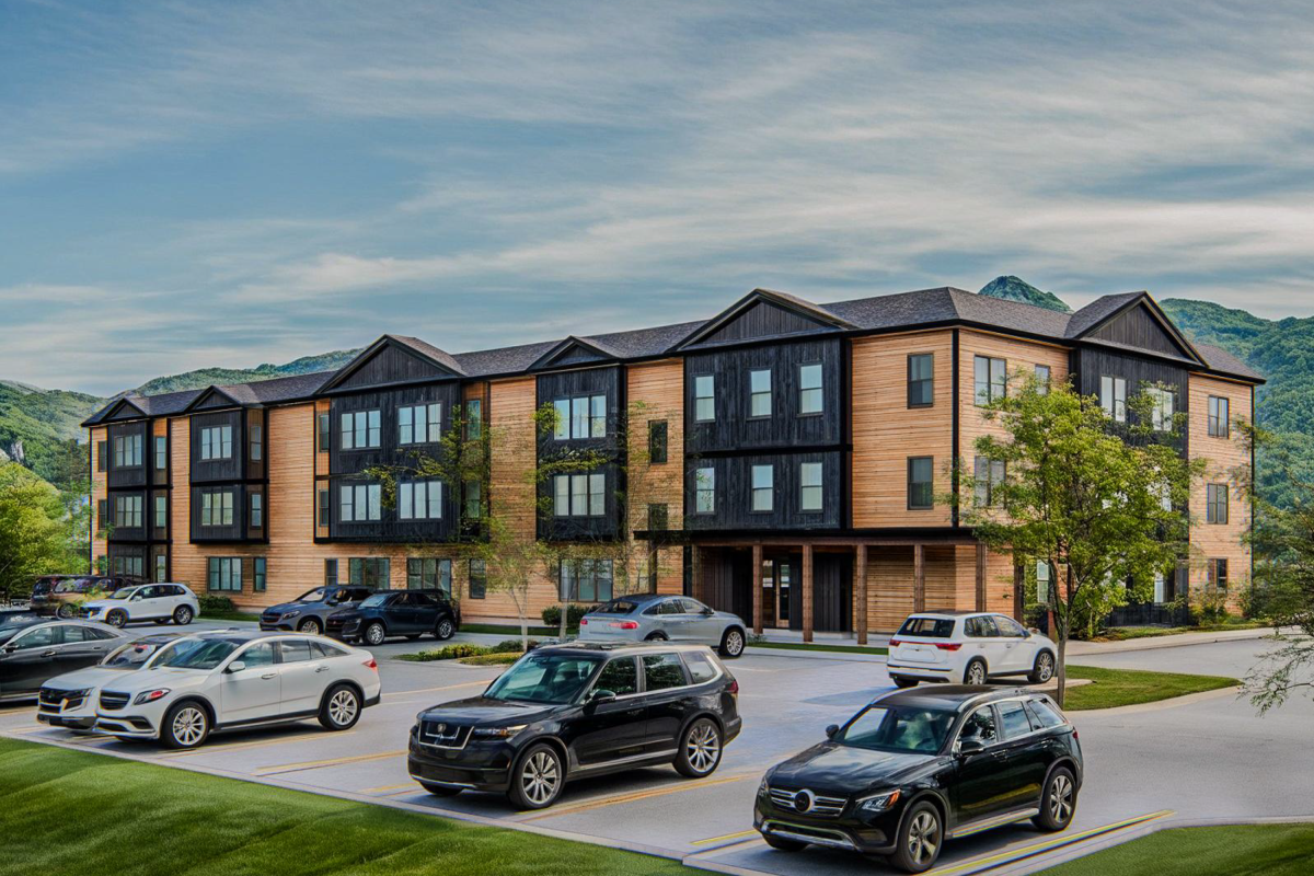 A three-story apartment building with wood and dark paneling, surrounded by parked cars and greenery, with mountains in the background under a cloudy sky.