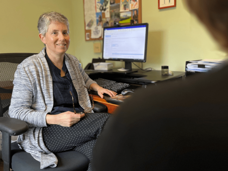 A person with short gray hair sits at a desk in an office, holding glasses, with a computer monitor and bulletin board in the background. Another figure is blurred in the foreground.