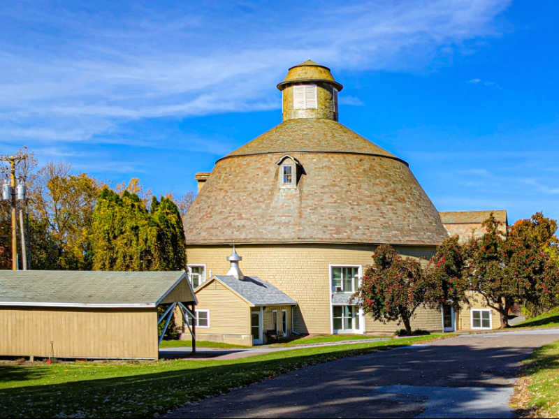 A round barn with a domed roof set in a rural landscape. Surrounding trees have autumn foliage. A small rectangular building is nearby under a clear blue sky.