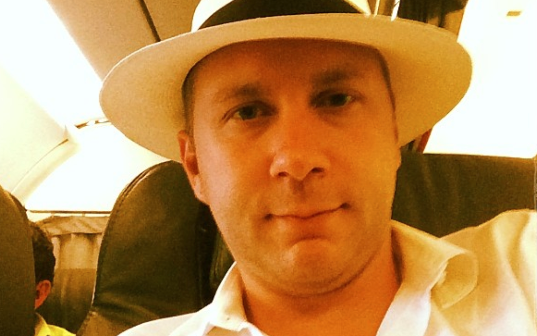 A man wearing a white hat and white shirt sits in an airplane seat, looking at the camera.